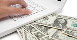 Hand typing on a white laptop keyboard with dollar bills underneath, representing how to make money online without paying anything.