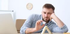 Man looking bored at a desk while holding a crumpled piece of paper near a laptop and notebook.
