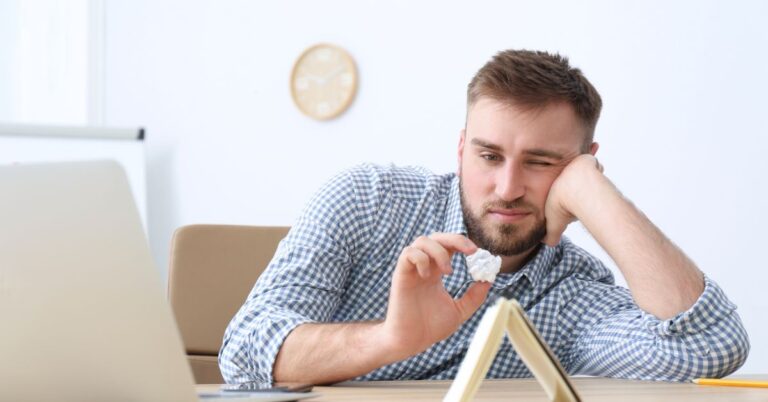 Man looking bored at a desk while holding a crumpled piece of paper near a laptop and notebook.