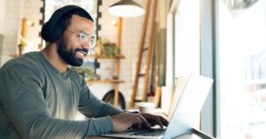 Man wearing headphones and glasses while writing on a laptop at a desk by a window.