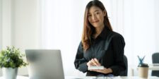 Woman seated at a desk with a laptop, holding a small notebook in a bright office.