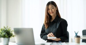 Woman seated at a desk with a laptop, holding a small notebook in a bright office.