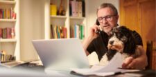 Small business owner working from home, talking on the phone while looking at a laptop with a dog on his lap and paperwork on the desk.