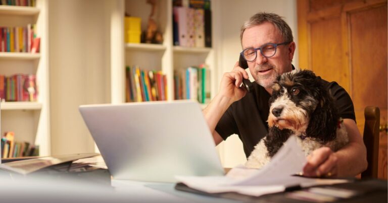 Small business owner working from home, talking on the phone while looking at a laptop with a dog on his lap and paperwork on the desk.