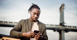 Man using a smartphone outdoors with a bridge in the background.