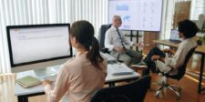 Three coworkers reviewing website or marketing information on computer screens during an office discussion.