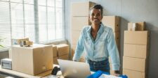 Small business owner standing behind a table with a laptop and packaging supplies, surrounded by stacked cardboard boxes in a bright workspace.