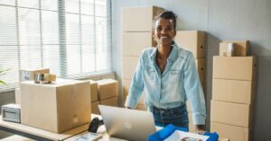 Small business owner standing behind a table with a laptop and packaging supplies, surrounded by stacked cardboard boxes in a bright workspace.