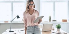 Woman holding a tablet while standing beside a desk with an open laptop in a bright office.