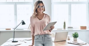 Woman holding a tablet while standing beside a desk with an open laptop in a bright office.