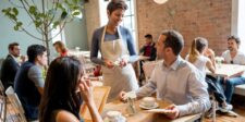 Server taking an order from customers in a busy café with people dining in the background.