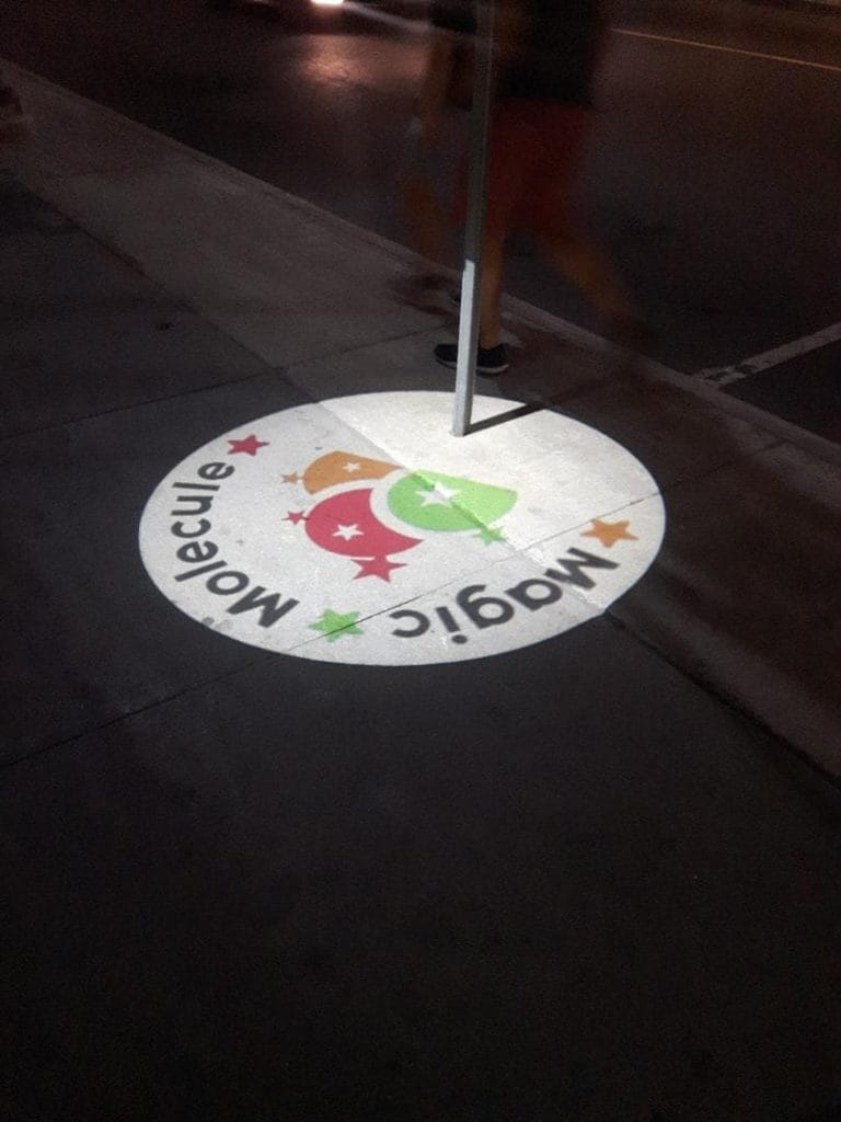 Circular “Magic Molecule” logo projected onto a sidewalk at night.