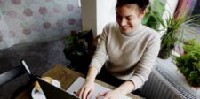 Woman smiling while working on a laptop at a café table beside a window.