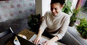 Woman smiling while working on a laptop at a café table beside a window.
