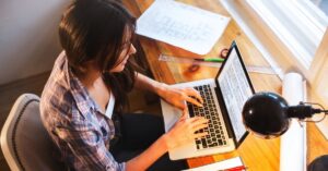 Woman working on a laptop at a wooden desk with papers, rulers, and a desk lamp nearby.