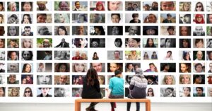 Three people sitting on a bench viewing a large wall display filled with photos of diverse faces.