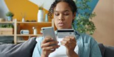 Shopify review (2026): Pros, cons, costs, and whether it's worth it Person shopping online with a smartphone and payment card while sitting on a couch at home.