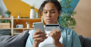 Person shopping online with a smartphone and payment card while sitting on a couch at home.