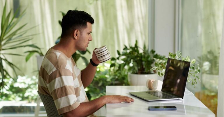Man sipping coffee while working on a laptop at a bright desk surrounded by indoor plants.