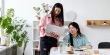 Two women collaborating in a bright office, with one holding a laptop while the other writes in a notebook at a desk.