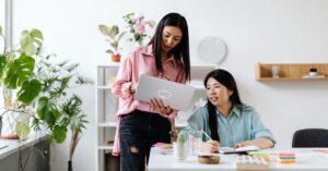 Two women collaborating in a bright office, with one holding a laptop while the other writes in a notebook at a desk.