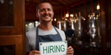Smiling small business owner holding a “Hiring” sign at the entrance to his business.