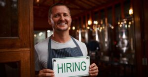 Smiling small business owner holding a “Hiring” sign at the entrance to his business.