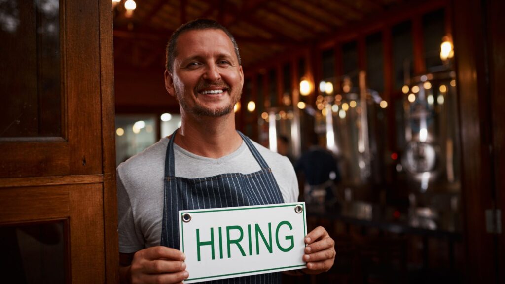 A small business owner standing with a sign that says "hiring."