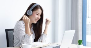 Woman wearing headphones smiling while using a laptop at a bright desk near a window.