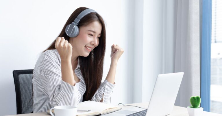 Woman wearing headphones smiling while using a laptop at a bright desk near a window.