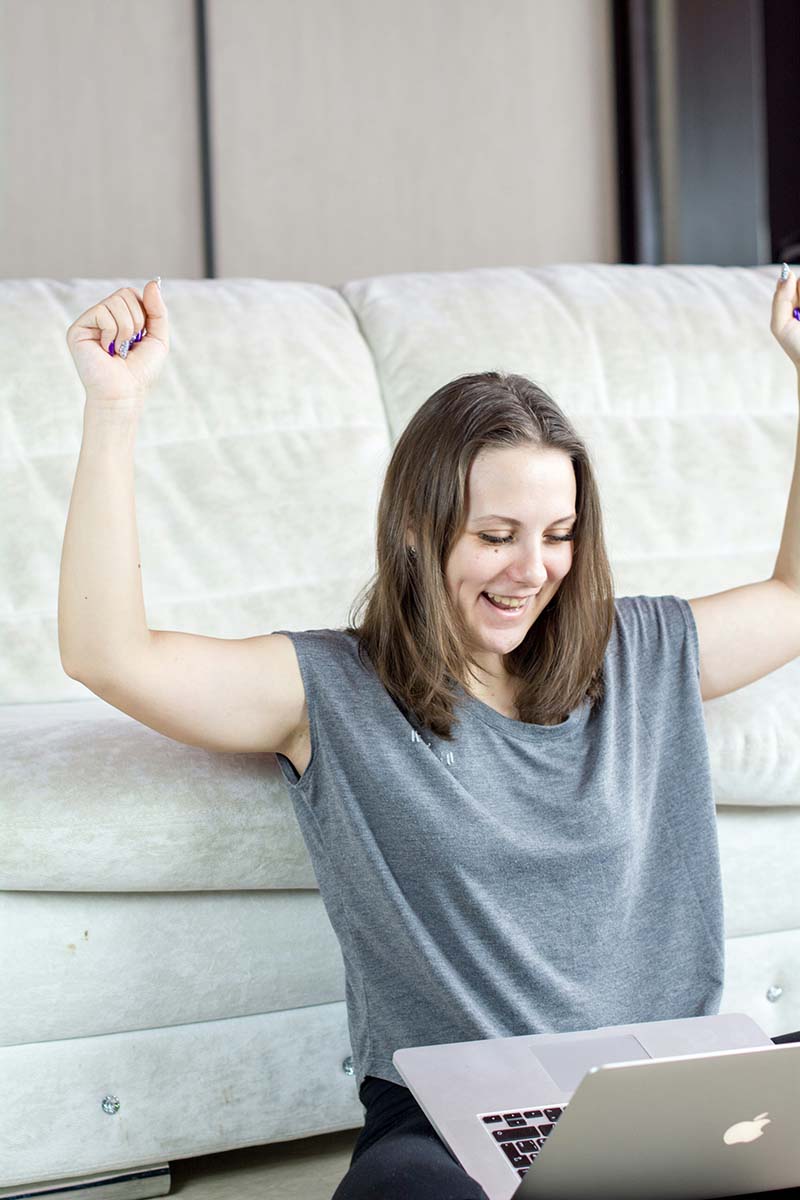 A woman sitting with a laptop on her lap, her arms raised in a victorious or celebratory gesture. She is smiling and looking at the laptop screen, suggesting she is happy with something she has seen or accomplished on the computer, maybe a domain name registration