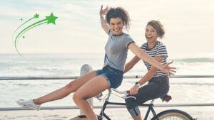 Two friends are enjoying a ride on a tandem bicycle by the beach, with smiles and arms raised in joy. The image captures the essence of how to live life with purpose, embracing moments of happiness and connection.