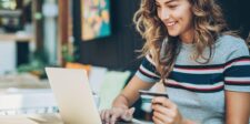 A smiling woman holds a credit card and types on her laptop in a cozy cafe, illustrating how to turn window shoppers into buyers.