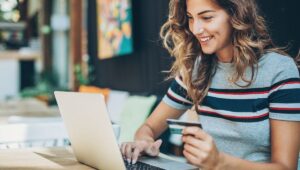 A smiling woman holds a credit card and types on her laptop in a cozy cafe, illustrating how to turn window shoppers into buyers.