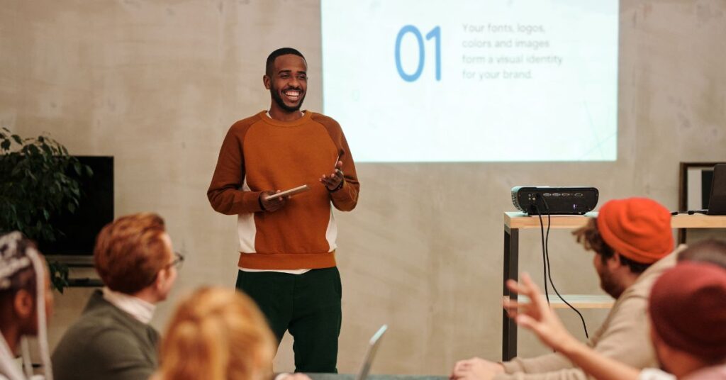 A confident speaker demonstrates how to work a room by engaging with his audience during a presentation. He is standing at the front of the room with a tablet in hand, smiling as he interacts with the seated attendees. A slide projected on the wall behind him displays the number '01' and text about visual identity for branding. The audience appears attentive and responsive, showcasing effective room-working skills.