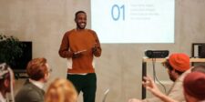 A confident speaker demonstrates how to work a room by engaging with his audience during a presentation. He is standing at the front of the room with a tablet in hand, smiling as he interacts with the seated attendees. A slide projected on the wall behind him displays the number '01' and text about visual identity for branding. The audience appears attentive and responsive, showcasing effective room-working skills.