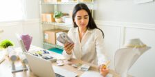 A young woman sits at a desk in a small office or workspace, holding a stack of cash while reviewing financial documents. She is surrounded by jewelry, including necklaces and earrings, suggesting she may be running a small business. A laptop and various tools are also on the desk, indicating she is managing her business operations. Shelving with boxes and more jewelry displays are visible in the background. The scene conveys a sense of financial management and entrepreneurial growth.