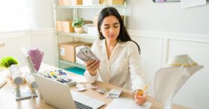 A young woman sits at a desk in a small office or workspace, holding a stack of cash while reviewing financial documents. She is surrounded by jewelry, including necklaces and earrings, suggesting she may be running a small business. A laptop and various tools are also on the desk, indicating she is managing her business operations. Shelving with boxes and more jewelry displays are visible in the background. The scene conveys a sense of financial management and entrepreneurial growth.