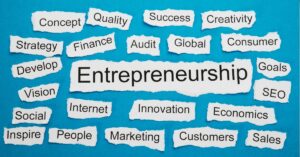 A graphic featuring the word "Entrepreneurship" prominently displayed in the center, surrounded by various related terms such as "Strategy," "Finance," "Innovation," "Marketing," "Success," and "Creativity." Each word is written on torn pieces of paper arranged against a blue background. The overall design conveys the diverse aspects of entrepreneurship and the resources needed to unlock success in this field.