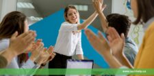 A woman in a white shirt smiles while giving a high-five to a colleague during a meeting, symbolizing teamwork and successful collaboration. Other team members are clapping, showing support and recognition for the achievement. A laptop is visible on the table, indicating a productive work environment. The image reflects themes of entrepreneurship and leadership, highlighting the importance of fostering a supportive and motivated team.