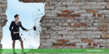 A woman in a business suit uses a sledgehammer to break through a brick wall, symbolizing how to overcome adversity by breaking down barriers and challenges.