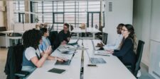 A diverse group of professionals engaged in a serious discussion around a conference table in a modern office setting, with laptops and documents in front of them.