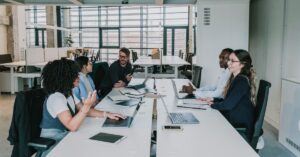 A diverse group of professionals engaged in a serious discussion around a conference table in a modern office setting, with laptops and documents in front of them.