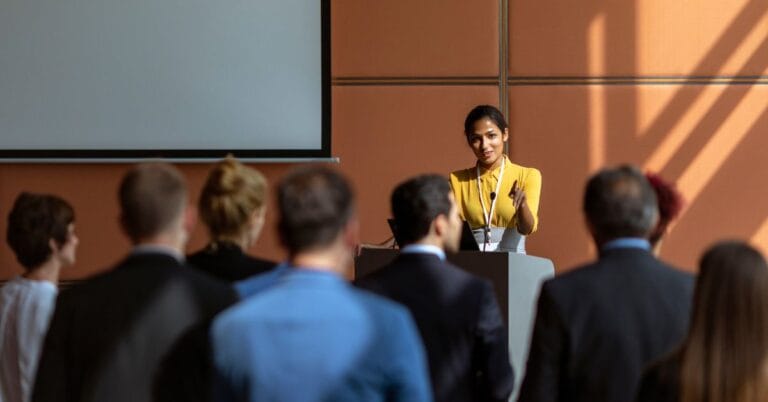 A woman confidently delivers a presentation to a group of attentive professionals, standing at a podium in a well-lit conference room. With poised body language and purposeful gestures, she effectively demonstrates the art of owning a room, engaging her audience with authority and presence.