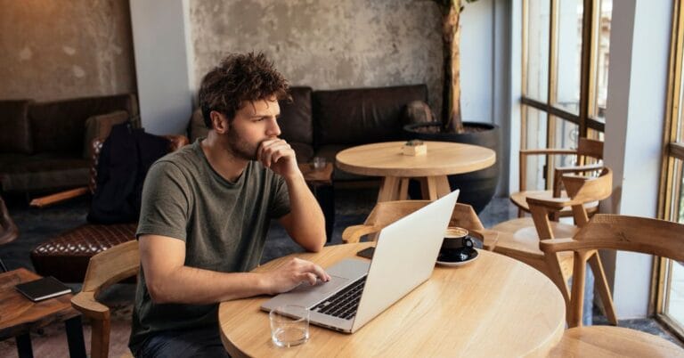 Man thoughtfully working on a laptop in a cozy cafe setting, symbolizing focus and reflection on important financial lessons for freelancers and entrepreneurs.