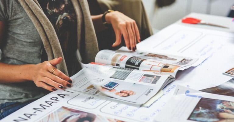 Person reviewing a magazine, engaging with various advertisements, while sitting at a desk filled with papers, scissors, and other creative tools.