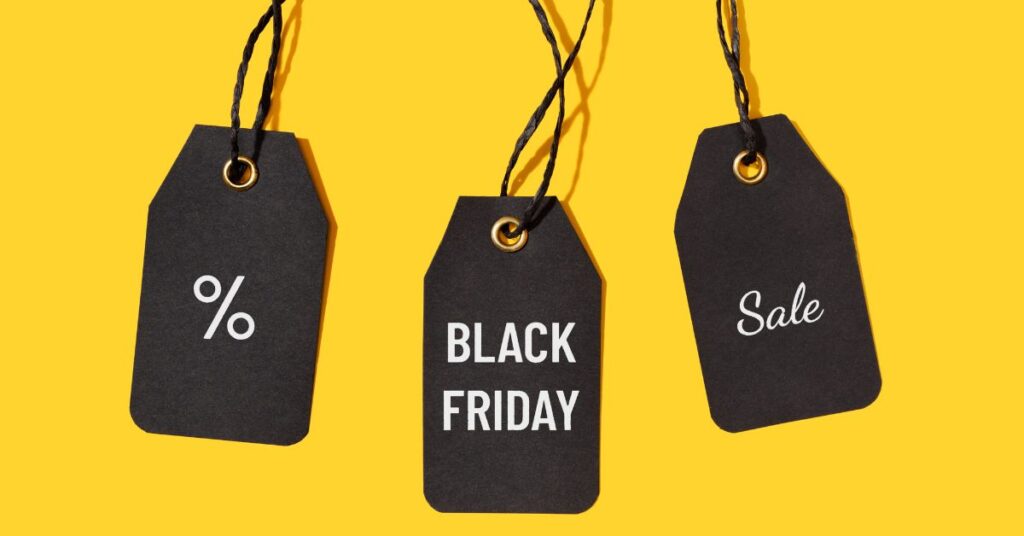 Three black price tags hanging against a bright yellow background. The tags display symbols and text: one has a percentage symbol (%), the middle reads 'Black Friday,' and the third says 'Sale,' highlighting discounts for a holiday sale