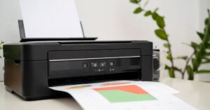 Close-up of a black inkjet printer with paper being printed featuring colorful charts, set against a background of green plants. The image illustrates one of the types of printing for home or office use, highlighting the use of color ink technology in producing documents and graphics.