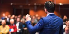 A speaker in a blue suit addresses an attentive audience during a presentation, illustrating the fundamentals of public speaking, including engaging delivery and audience connection.