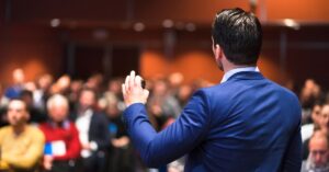 A speaker in a blue suit addresses an attentive audience during a presentation, illustrating the fundamentals of public speaking, including engaging delivery and audience connection.